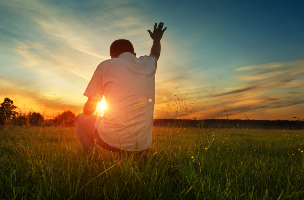 A person kneeling in a field with one hand raised towards the sky during a sunset, symbolizing an open heart to God.