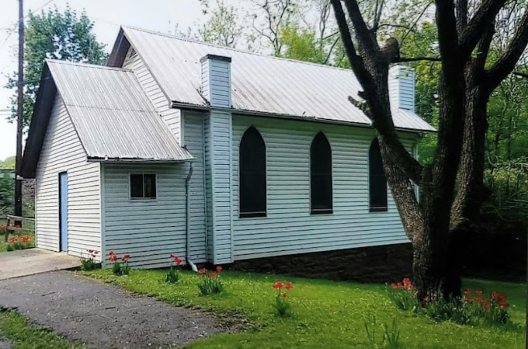 Exterior of Little Falls Chapel, a historic white church with arched windows, surrounded by greenery and flowers.