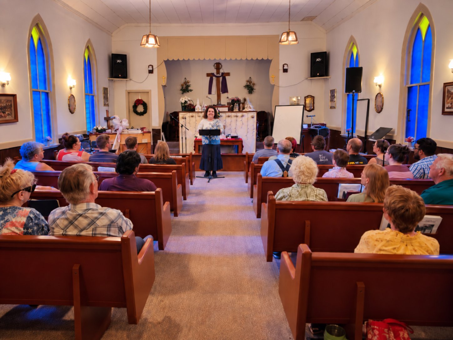 Sunday worship at Little Falls Chapel: Pastor Linda Reese speaking to the congregation seated in wooden pews beneath blue arched windows